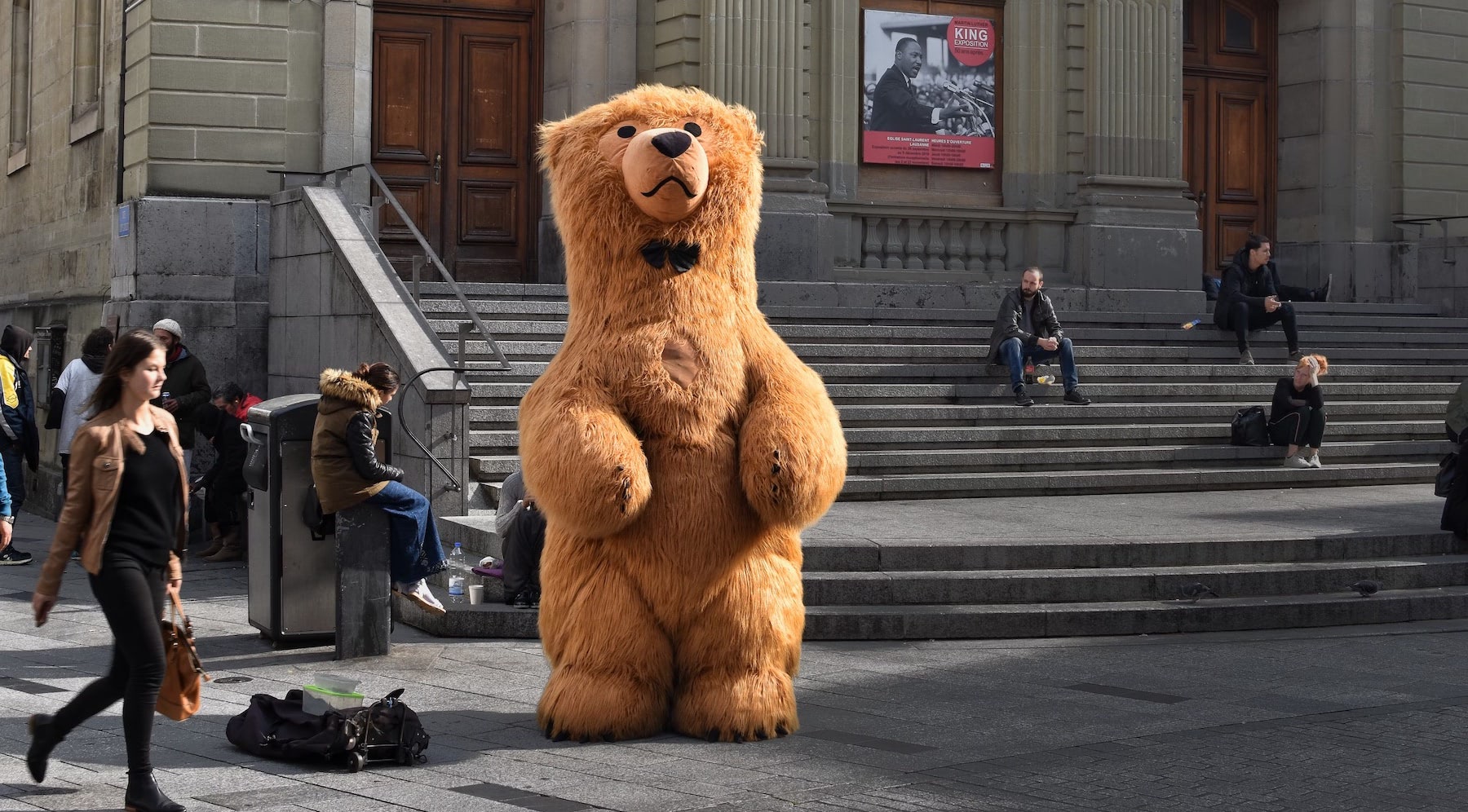 A person wearing a brown fur coat stands in the street, holding a teddy bear in front of a tall outdoor building.